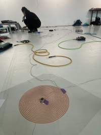 In the backgroud, a person dressed in black is squatting in front of a sound deck and a computer, which they are using. They are holding a microphone to their mouth. At the front, a red coil spiral is stuck to the floor. A thin cable is attached to its center with purple table. Throughout the rest of the space, other sound equipment and colourful cables lie on the floor.