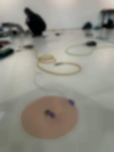 In the backgroud, a person dressed in black is squatting in front of a sound deck and a computer, which they are using. They are holding a microphone to their mouth. At the front, a red coil spiral is stuck to the floor. A thin cable is attached to its center with purple table. Throughout the rest of the space, other sound equipment and colourful cables lie on the floor.