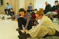 A group of people is sitting on the floor or on chairs. They are all facing the same direction. At the front, two people are sitting close together. One of them points to something on the floor, and the other, looking in that direction, holds a microphone to their mouth.