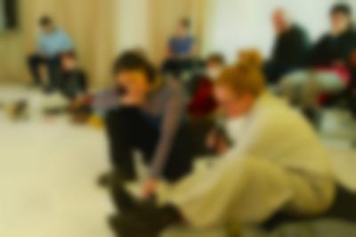 A group of people is sitting on the floor or on chairs. They are all facing the same direction. At the front, two people are sitting close together. One of them points to something on the floor, and the other, looking in that direction, holds a microphone to their mouth.