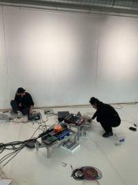 Decks and other sound equipment have been set in two low tables and on the floor. A person can be seen squatting next to the larger structure, using a laptop. Someone else is sat by the wall, with a laptop to the left and a deck to the right, which they are adjusting.