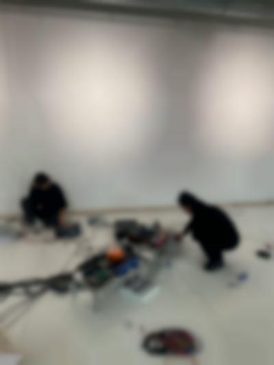 Decks and other sound equipment have been set in two low tables and on the floor. A person can be seen squatting next to the larger structure, using a laptop. Someone else is sat by the wall, with a laptop to the left and a deck to the right, which they are adjusting.