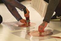 Close-up of two coil spirals, stuck to the white floor with a transparent plastic. There are wires coming from each of these spirals, and another wire connecting them both. The arms and a foot of two people can be seen. One person holds a microphone to a magnet, that they have set on one of the spirals. The other person touches the centre of the other spiral.