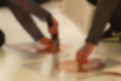 Close-up of two coil spirals, stuck to the white floor with a transparent plastic. There are wires coming from each of these spirals, and another wire connecting them both. The arms and a foot of two people can be seen. One person holds a microphone to a magnet, that they have set on one of the spirals. The other person touches the centre of the other spiral.