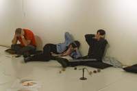 Three people are sitting or lying on black cushions on the floor, next to a white wall. They are sitting in different positions, but are all holding their hands to their ears.