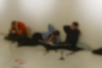 Three people are sitting or lying on black cushions on the floor, next to a white wall. They are sitting in different positions, but are all holding their hands to their ears.