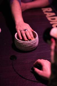 A hand hovers over a small basket, possibly made from cotton or similar material. The basket is set on a dark table. The hand of someone else, sitting across, rests on the table.