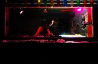 A person stands in a dark DJ booth, adjusting the knobs on a DJ deck. To their right, a bright light shines upwards from a light box that is set next to the deck.