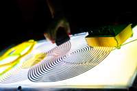 On top of a light box, there are two coil spirals and a small sound deck with knobs. A hand can be seen holding an object, placed on top of one of the spirals.