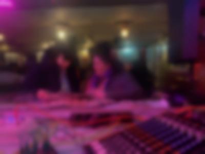 Next to a sound deck, two people are looking through a newspaper and smiling. They are in an indoor space after dark, possibly a bar or a cafe.