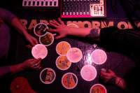 A number of round tarot cards have been set with no apparent order on top of a dark table. Someone is holding a card with both hands, while the person sitting across from them appears to be touching or picking a card. Next to the cards, two sound decks are partially visible.