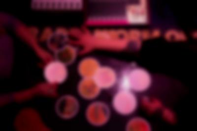 A number of round tarot cards have been set with no apparent order on top of a dark table. Someone is holding a card with both hands, while the person sitting across from them appears to be touching or picking a card. Next to the cards, two sound decks are partially visible.