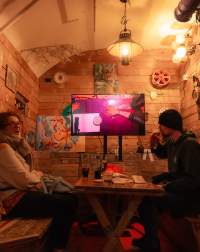 Two people sit across from each other. In between them there is a table with drinks and paper materials. One of them takes a sip from a beer. At the back, there is a screen on a stand, showing a table filmed from above. In this image, someone rests their arms on the table, with their palms facing up. Someone is sitting across from them. There is a sheet of paper and a stack of round tarot cards.