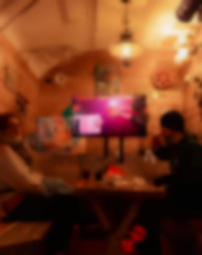 Two people sit across from each other. In between them there is a table with drinks and paper materials. One of them takes a sip from a beer. At the back, there is a screen on a stand, showing a table filmed from above. In this image, someone rests their arms on the table, with their palms facing up. Someone is sitting across from them. There is a sheet of paper and a stack of round tarot cards.