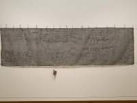 A large rectangular piece of fabric has been hung on the wall. There is some texture to it, created with stitching. At the bottom, one small object hangs from the fabric.