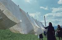 Two feminine-presenting people, dressed in dark colours and wearing head scarves, stand by a clothesline full with white fabric in a natural setting.