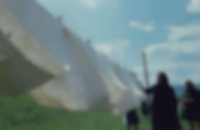 Two feminine-presenting people, dressed in dark colours and wearing head scarves, stand by a clothesline full with white fabric in a natural setting.