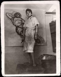 The artist is standing in front of a nude study. She has short hair and is wearing a wrapped white coat, which is smudged with charcoal.
