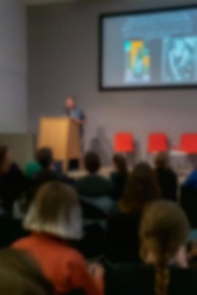 Nearly full lecture room. On stage, a person stands behind a podium. There is slide being projected behind them. It has a text, a painting and a photograph in black-and-white.