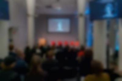 Nearly full lecture room. On stage, a person stands behind a podium. There is an image being projected on stage and in another screen, which is closer to the middle of the room. The image shows a feminine-presenting person with short hair in black-and-white.