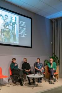Four people sit on stage, around a low round table. A slide with a photograph and a short text is being projected behind them. The person to the left holds a microphone.