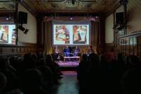Event room with several rows of chairs full of people. In front of them, four people side around a low table on stage. A slide with two photographs is being projected above them.