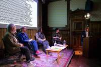 Four people sit around a low table. To their right, another person is behind a podium, towards which the others are looking. A slide with the tile of the book, 