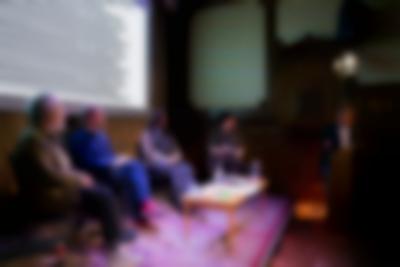 Four people sit around a low table. To their right, another person is behind a podium, towards which the others are looking. A slide with the tile of the book, 