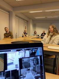 A laptop screen is partially visible, with several  photo files open on the desktop. Behind it, five people sit around tables in a university room.