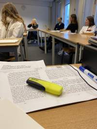 Two text print outs have been set on a table. A thick yellow highlighter lays on top. In the background, five other people sit around other tables, looking in the same direction.
