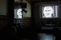 A film is being projected on a screen. The image shows a head shot of a masculine-presenting person with a moustache. The image also appears partly reflected on a mirror to the left.
