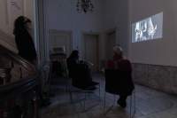 Three people watch a film being projected on a wall of a hallway space. Two of them are sitting down, while the other stands up at the bottom of the stairs.