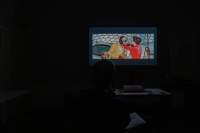 Dark space. Only the projection of a film is visible.  The image shows a feminine-presenting person touching the head of another person.