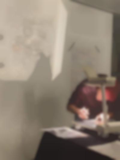 A person is leaning over an overhead projector. They are making notes on the sheet on the projector's stage. The image is being projected behind him. Next to the projection, a white sheet of paper with notes, handwritten in different colours, has been stuck to the wall with two thin strips of black tape.