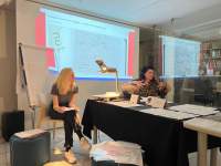 Two people sit in front of a projection and a flip chart easel. On a table, in front of them, there is an overhead projector and several sheets of paper and other printed material.