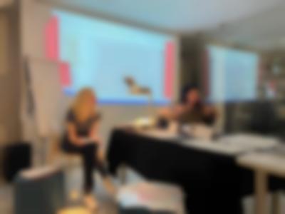 Two people sit in front of a projection and a flip chart easel. On a table, in front of them, there is an overhead projector and several sheets of paper and other printed material.