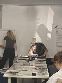 At the edge of a long table, on top of which there are several sheets of paper, a person is leaning over an overhead projector. They are making notes on the sheet on the projector's stage. Next to them, someone stands in front of a flip chart easel.