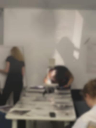 At the edge of a long table, on top of which there are several sheets of paper, a person is leaning over an overhead projector. They are making notes on the sheet on the projector's stage. Next to them, someone stands in front of a flip chart easel.