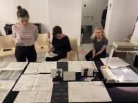 Three people are next to a table. Two are sitting and one is standing up. On the table, there are sheets of paper with handwritten notes and an overhead projector.