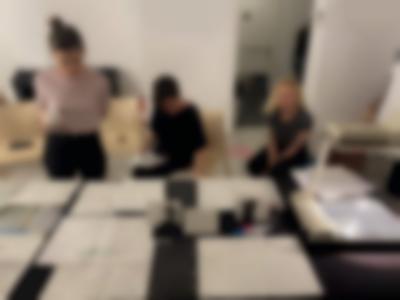 Three people are next to a table. Two are sitting and one is standing up. On the table, there are sheets of paper with handwritten notes and an overhead projector.