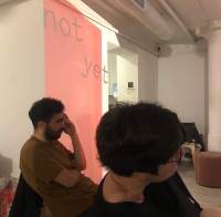 Two people are sitting, facing the same direction. Behind them, there is wall projection of a red block with the words 