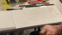 A white sheet of paper with notes and drawings is at the edge of a table. There are pens and pencils around it. The hand of the person sitting in front of the sheet can be see on the bottom right corner.