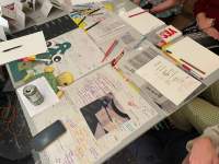 Several sheets with handwritten notes, printouts with notes and a photograph have been laid on a table. There are also pens and pencils, a smartphone and a can of beer. The laps and arms of three people, who are sitting around the table, are visible.
