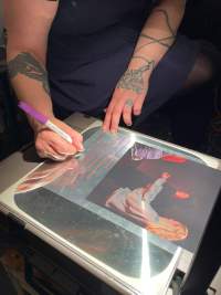 A person's hands and arms are partially visible. They are writing, with a purple marker, on a transparency sheet, under a photograph. The sheet is on an overhead projector's stage.