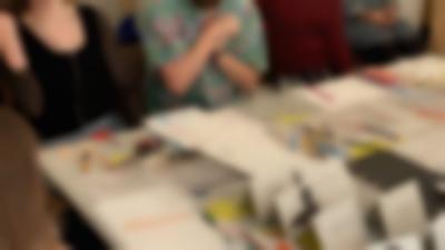 Several sheets with handwritten notes, printouts with notes and a photograph have been laid on a table. There are also pens and pencils, three glass and two scissors. Five people are partially visible around the table.