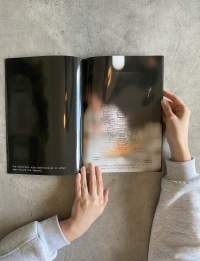 Two hands hold a book open. The left page is black, with white text at the bottom. The right page has a blurry image with a diagram, handwritten in black, on top. There is some text, written digitally in black, at the bottom.