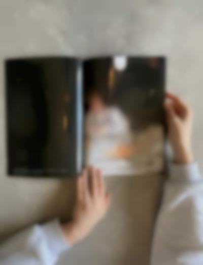 Two hands hold a book open. The left page is black, with white text at the bottom. The right page has a blurry image with a diagram, handwritten in black, on top. There is some text, written digitally in black, at the bottom.