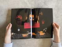 Two hands hold a book open. Across the two pages, there is a photograph of objects on top of a black background. A red lego is discernible in the middle. White text appears at the bottom, on both pages.