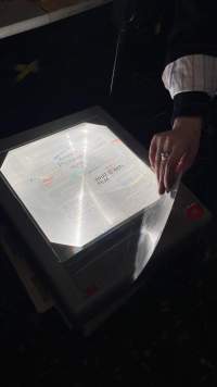 Handwritten notes, in several colours, have been written in a transparency sheet and left on top of an overhead projector's stage. A hand is partially visible, holding the corner of the sheet.