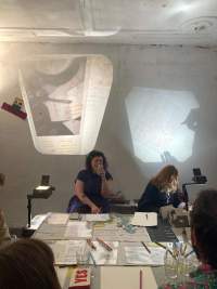 Four people sit around a table, on top of which there is printed material and pens and pencils. There two overhead projectors, one in each side of the table, projecting images to a wall. One of the people is writing on a sheet of paper, whose image is being projected on the wall.